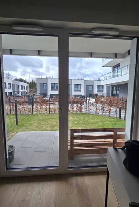 Widok z pokoju przez okno balkonowe oklejone folią Titan 25 Duo Premium na przyblokowy ogródek i nowoczesne osiedle. Folia nadaje szybie neutralny, lekko szarawy odcień, zapewniając komfort wizualny bez drastycznego zaciemnienia obrazu.